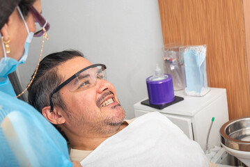A cheerful male patient wearing safety glasses smiles and talks with a masked dentist during a...
