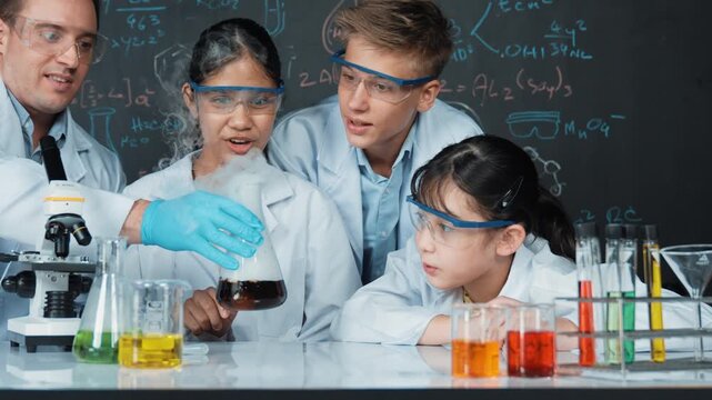 Girl looking at beaker while doing experiment at blackboard with chemistry theory in STEM class. teacher and group of high school student with mixed races doing science activity together. Edification