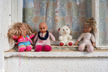 Four well-loved dolls, worn and expressive, sit on an old concrete windowsill against faded curtains. A nostalgic glimpse into childhood memories and timeworn toys.