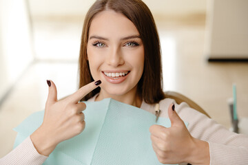 Smiling beautiful girl is sitting in dentistry chair with positive emotion and gesturing fingers...