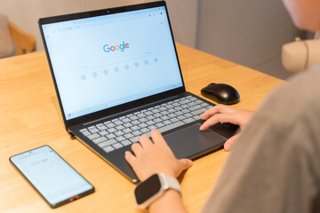 Person using laptop with Google search page open, smartphone and smartwatch on wooden desk, technology concept
