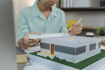Asian male architect sitting at a desk, measuring a modern house model with triangular ruler and pencil. Office setup includes design tools, blueprints, and safety gear, suggesting architectural work.
