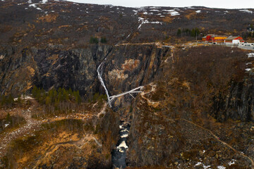 Magnificent Voringsfossen waterfall plunges into deep rugged canyon flanked by rocky cliffs. Winding road and sparse vegetation complete landscape