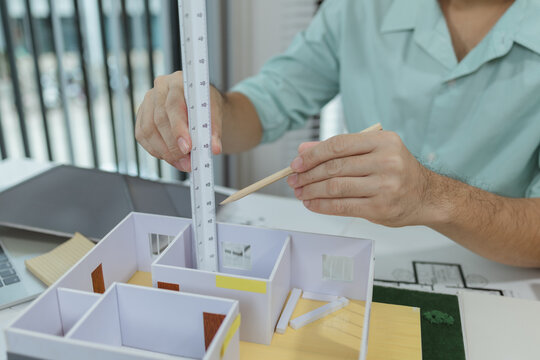 Asian male architect sitting at a desk, measuring a modern house model with triangular ruler and pencil. Office setup includes design tools, blueprints, and safety gear, suggesting architectural work.