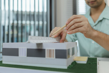 Asian male architect sitting at a desk, measuring a modern house model with triangular ruler and...
