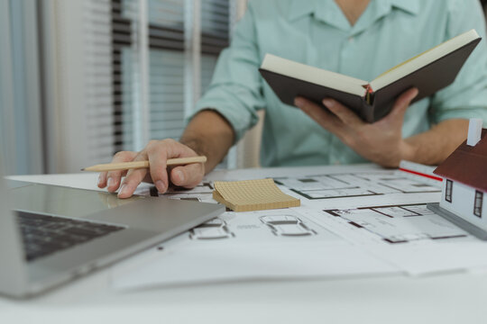 Architect working on a house plan using a pencil while referencing a notebook and laptop. A small house model and floor plan are placed on the desk, showing a detailed architectural layout.