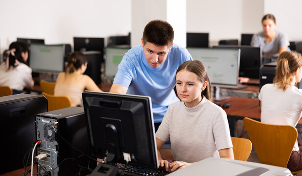 Smart interested teen schoolboy helping cute girl classmate sitting at computer during informatics lesson