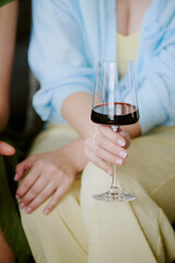 Woman holding wine glass with red wine, sitting with another partially visible, showing manicured hands and relaxed posture, focus on hand and drink
