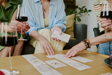 Group of middle aged Caucasian women sitting together holding glasses of red wine and arranging tarot cards on table, hands and arms visible, engaging in fortune telling activity