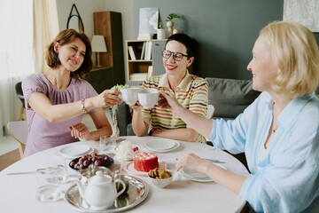 Three middle aged Caucasian women sitting at table clinking cups and smiling, enjoying conversation together, sharing dessert and fruit, celebrating friendship in home dining room