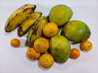 A mix of fresh fruits, including green mangoes, ripe yellow bananas, and multiple small oranges. Clustered on a white surface, stressing their varying colors and textures, relating to tropical fruits
