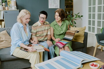 Three middle aged Caucasian women sitting on sofa discussing fabric swatches and interior design samples, holding textile samples and looking at open catalogs together