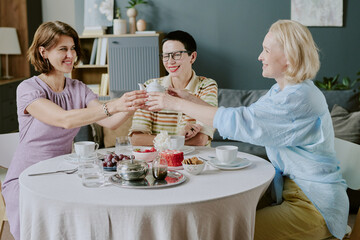 Three middle aged women sitting at round table clinking cups, smiling and enjoying conversation during breakfast with fruit and pastries