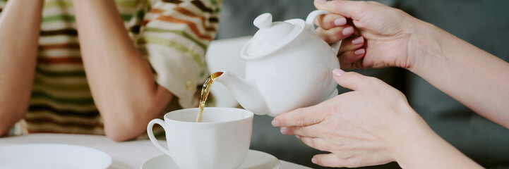 Unrecognizable woman pouring tea from ceramic teapot into cup while sitting at table with another...