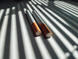 Two cigarettes with an orange filter lie on a white windowsill in the shadow of the blinds, which...