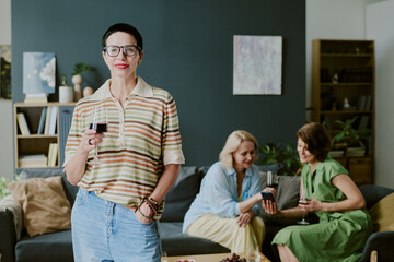 Obraz premium Portrait of middle aged Caucasian woman with short hair and glasses holding wine glass, standing in living room while her girlfriends sitting on sofa talking and drinking wine