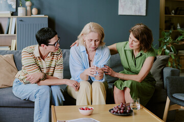 Middle aged Caucasian woman sitting on sofa holding glass of water looking down, supported by two caring girlfriends comforting her with gentle touch, bowl of fruit on table