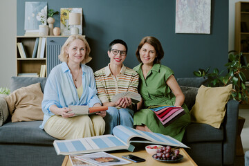 Three middle aged Caucasian women sitting on sofa smiling and holding fabric swatches, engaging in interior design discussion with open catalogs and digital tablet on table