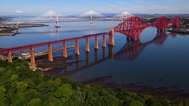 Established aerial video of a train travelling over the Forth Bridge in Scotland, with Forth Road Bridge and Queensferry Crossing on the background. 