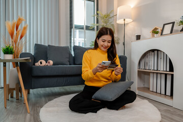 Young Asian woman sitting cross-legged on a rug at home, holding a credit card and smartphone. She...