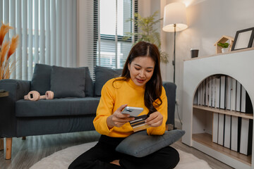 Young Asian woman sitting cross-legged on a rug at home, holding a credit card and smartphone. She...