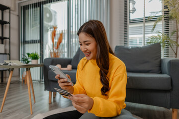 Young Asian woman sitting cross-legged on a rug at home, holding a credit card and smartphone. She...