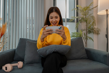 Young Asian woman in yellow sweatshirt sitting on sofa at home, intensely focused on playing mobile...