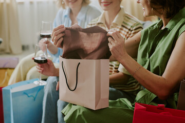 Three Caucasian middle aged women sitting together holding wine glasses and shopping bags, one woman smiling while taking clothing item out of gift bag during social gathering