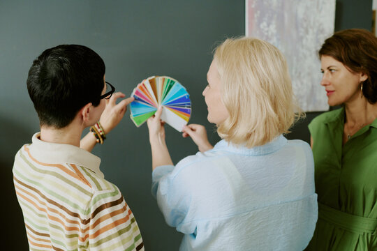 Three middle aged women standing together examining color swatch samples against dark wall, discussing color choices - Powered by Adobe