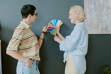 Middle aged Caucasian woman showing color swatches to another woman, both standing near wall and...