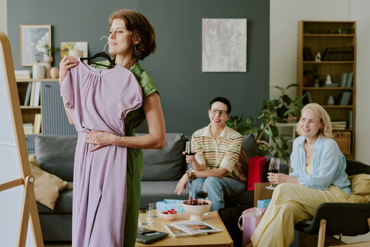 Middle aged Caucasian woman holding dress in front of mirror while two female friends sitting on sofa watching and smiling during casual gathering