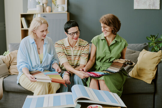 Three middle aged Caucasian women sitting on sofa examining fabric swatches together, smiling and discussing textile samples, large open sample books with striped patterns on table - Powered by Adobe