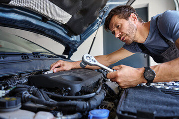 Male mechanic working on car engine with wrench under open hood in garage. Man in work overalls is...