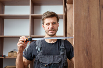 Man in work overalls holding measuring tape during assembling wooden wardrobe. Concept of DIY home...