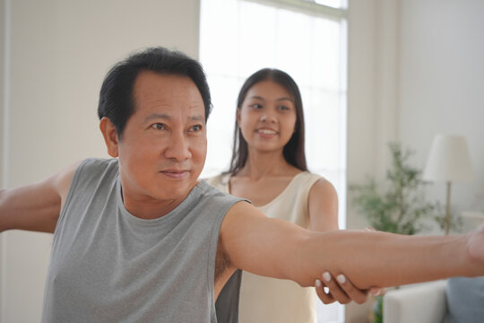 Asian Middle aged man getting support from young woman while stretching arms during home yoga session in a room