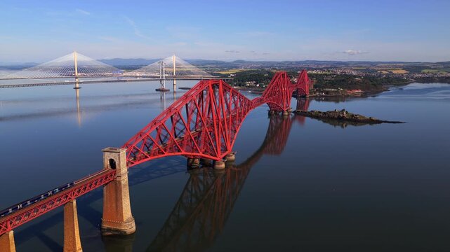 Aerial video of a train travelling over the Forth Bridge in Scotland, with Forth Road Bridge and Queensferry Crossing on the background. 