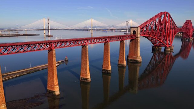Aerial video of a train travelling over the Forth Bridge in Scotland, with Forth Road Bridge and Queensferry Crossing on the background. 