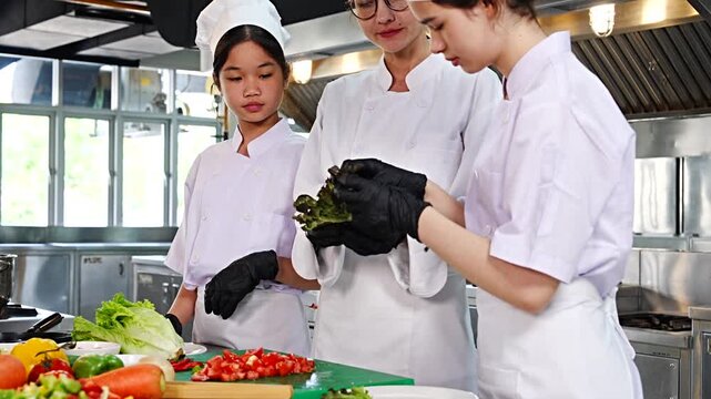 Female chef teaches cooking to two teenage girls, learning how to select vegetables, preserve food and cook, everyone wears black gloves in cooking class.