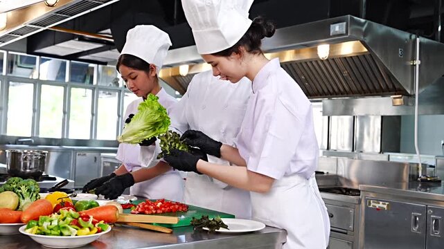Female chef teaches cooking to two teenage girls, learning how to select vegetables, preserve food and cook, everyone wears black gloves in cooking class.