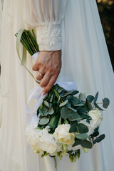 A bride in a long white wedding dress embroidered with various patterns holds a bouquet of white...