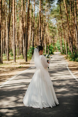 The bride in a white long wedding dress with her back to the camera stands on an asphalt road in...
