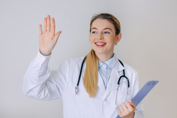 Smiling doctor waving while holding clipboard in bright clinic office setting