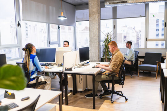 Diverse team of young professionals concentrating on their computer screens in a bright, modern coworking office, demonstrating teamwork and productivity in a shared workspace