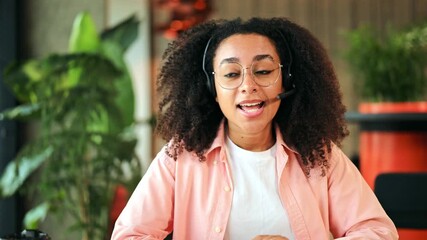 Cheerful African American woman wearing a headset and pink shirt actively engages in an online video call from a bright modern office. She provides excellent customer support with a friendly smile.
