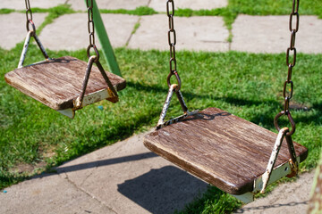 Paired weathered wooden swing seats hanging from corroded metal chains above green grass,...