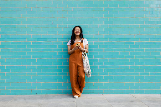 Smiling person with reusable shopping bag using smartphone by turquoise brick wall