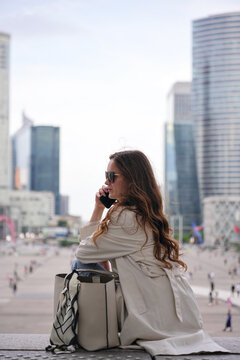 A woman talks on her smartphone while sitting in La Defense business district in Paris with modern skyscrapers and an urban backdrop 