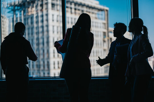 Silhouettes of a diverse group of professionals in a city building discussing aspects of construction or architecture while a bustling urban skyline provides an inspiring backdrop.