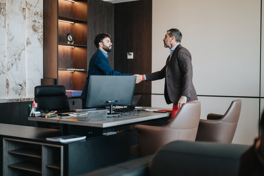 Two business individuals shaking hands in a professional office, showcasing agreement or partnership, with an elegant and contemporary interior design in the background.