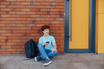 Cheerful Redhead Teen Boy Texting on Phone Outside School Wall